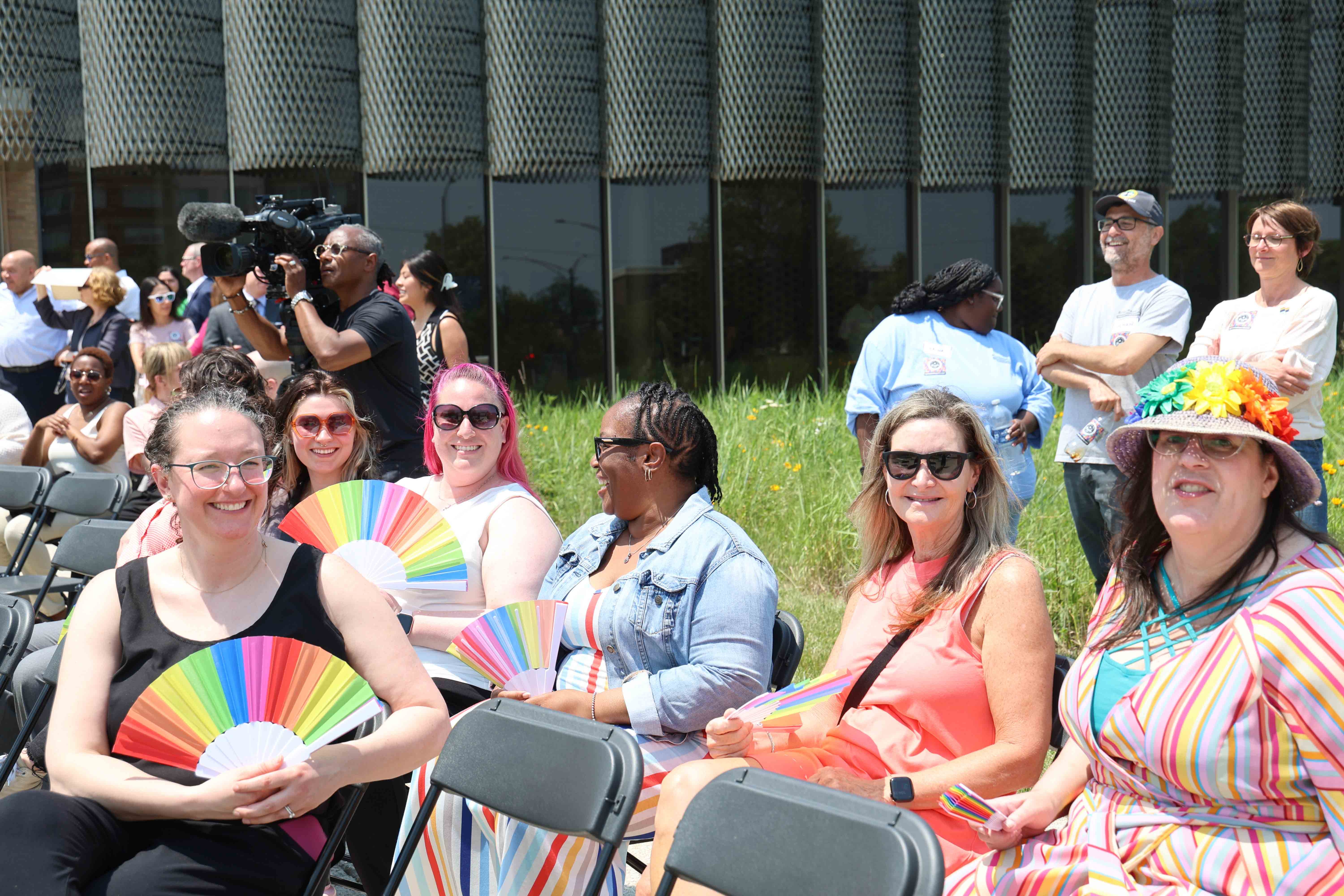 People with rainbow fans sit outdoors at a Pride event. A videographer films the crowd.